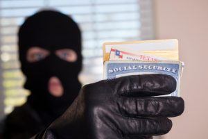 Masked theif wearing all black steals a victim's identification cards. Social security card, Texas driver's license, credit cards are being held in the criminal's black glove. Window background. Identity theft concept.